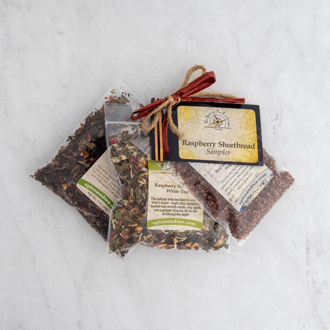 Three clear plastic bags of loose Raspberry Shortbread White Tea, bundled with a brown ribbon and a “Raspberry Shortbread Sampler” label, displayed on a light marble surface.