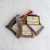 Three clear plastic bags of loose Raspberry Shortbread White Tea, bundled with a brown ribbon and a “Raspberry Shortbread Sampler” label, displayed on a light marble surface.