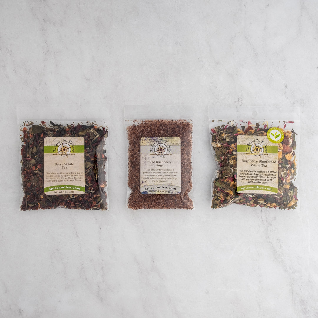 Three clear bags of loose leaf tea labeled Berry White Tea, Red Raspberry Sugar, and Raspberry Shortbread White Tea from the Raspberry Shortbread Sampler are displayed side by side on a light marble surface.
