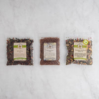 Three clear bags of loose leaf tea labeled Berry White Tea, Red Raspberry Sugar, and Raspberry Shortbread White Tea from the Raspberry Shortbread Sampler are displayed side by side on a light marble surface.