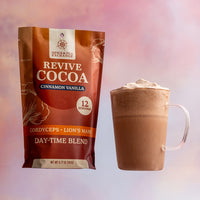 A bag of Revive Cocoa Cinnamon Vanilla and a glass mug of frothy cocoa sit against a soft pink and purple gradient background.