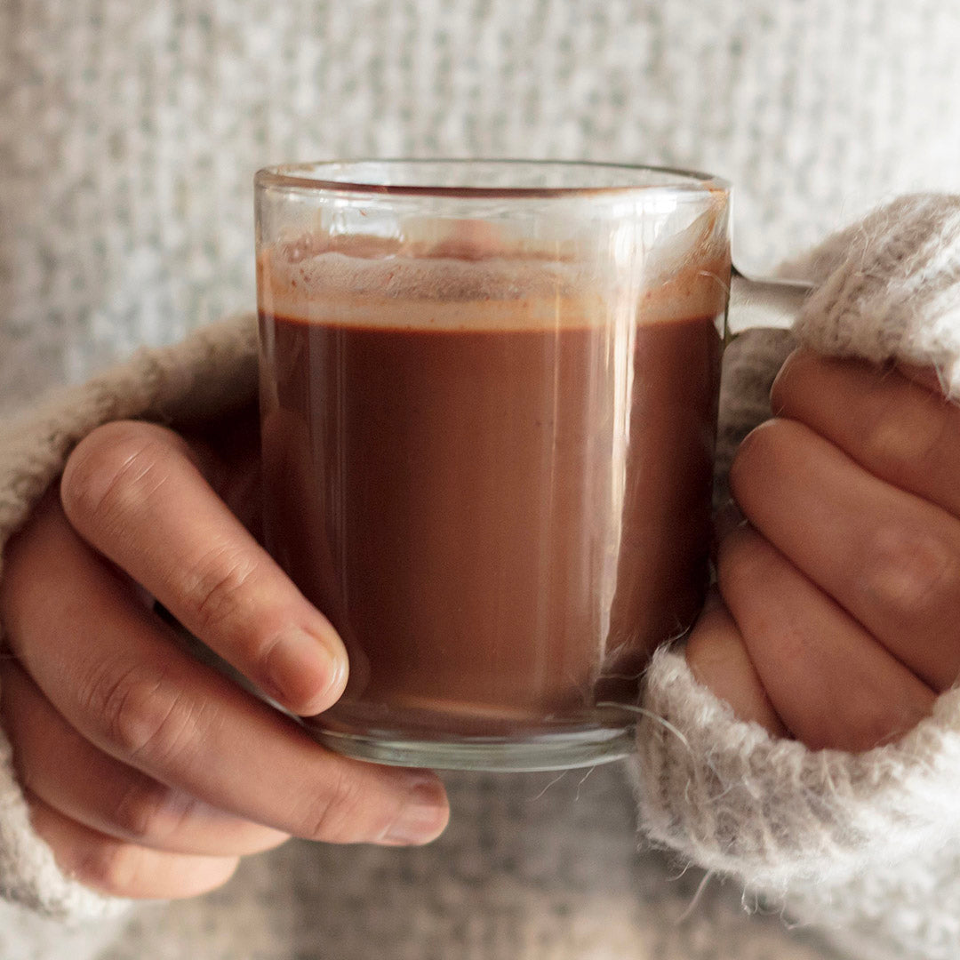 Glass mug of hot chocolate held by hands in cozy gloves against a neutral background