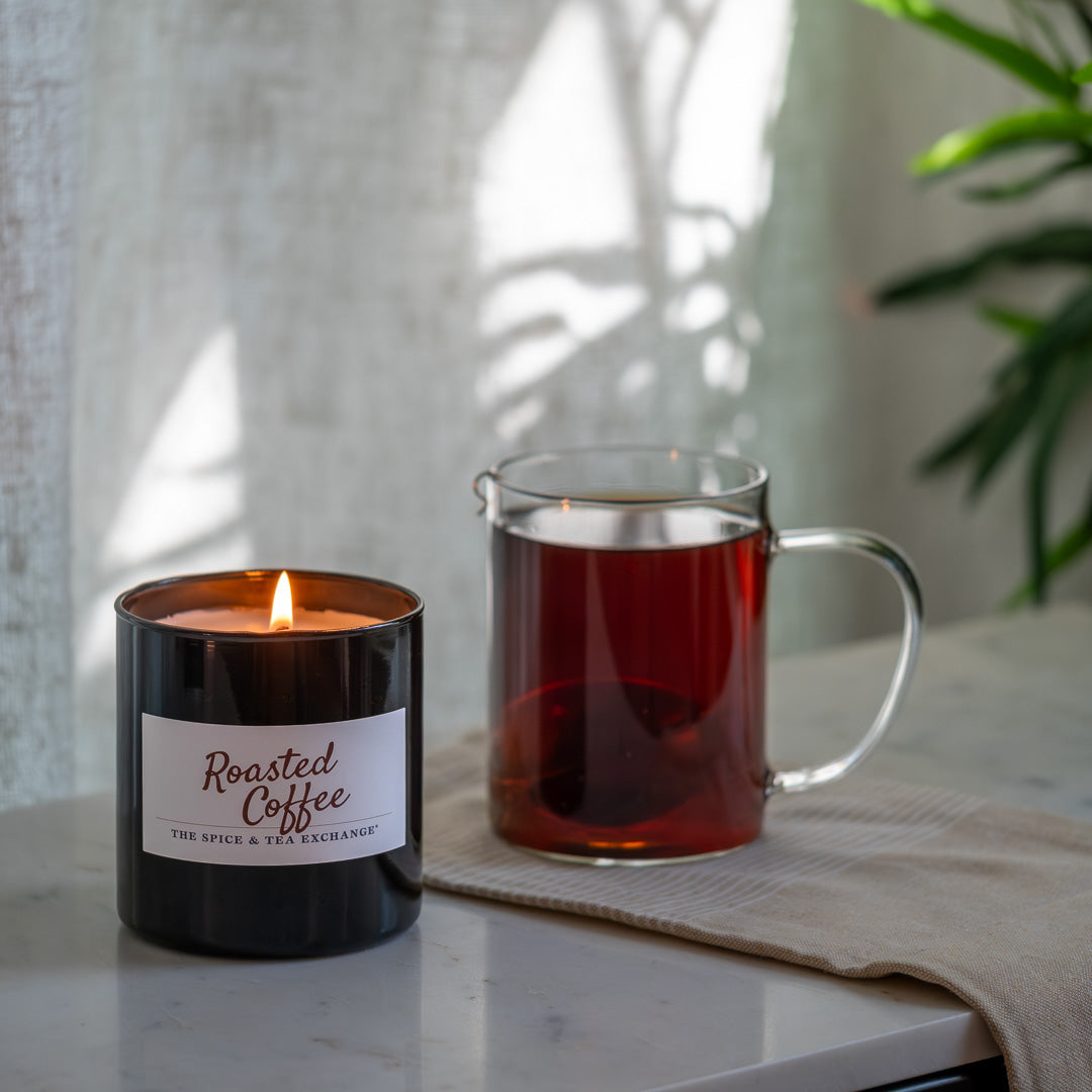 Candle labeled 'Roasted Coffee' next to a mug of tea on a surface with a plant in the background.
