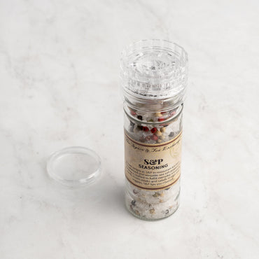 A clear plastic S & P Seasoning grinder, filled with mixed peppercorns and sea salt, sits on a light marble surface with its lid removed and placed beside it.