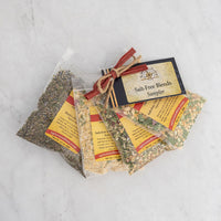 Four clear bags of assorted Salt-Free Blends Sampler spices are displayed on a white surface. Each bag features a yellow label, and a tag reading “Salt-Free Blends Sampler” is tied with twine and a red ribbon for healthy seasoning.