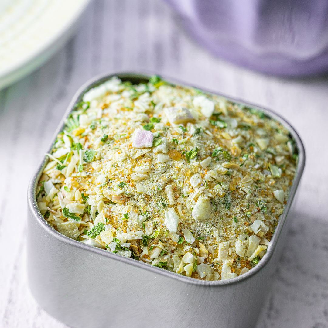 A close-up of the Salt-Free Onion Obsession Spice Blend, a metal container filled with a vibrant onion mix of dried herbs, spices, and chopped vegetables, ideal for creating salt-free dips and spreads on a light surface.