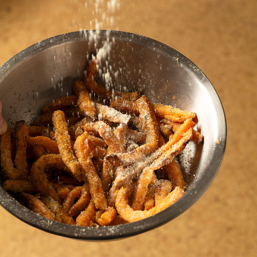 A metal bowl of crispy sweet potato fries is being sprinkled with French Fry Seasoning - Salt 'n Vinegar from above, with a blurred background highlighting the fries and the zesty seasoning as it falls.