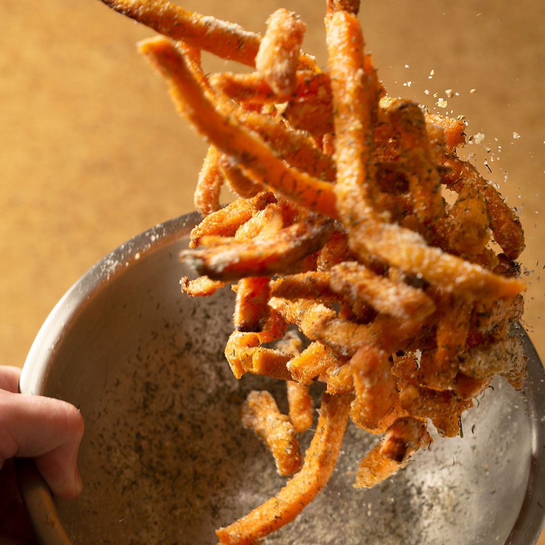 A close-up of sweet potato fries being tossed in French Fry Seasoning - Salt 'n Vinegar in a metal bowl, with flavorful seasoning particles visible in the air.