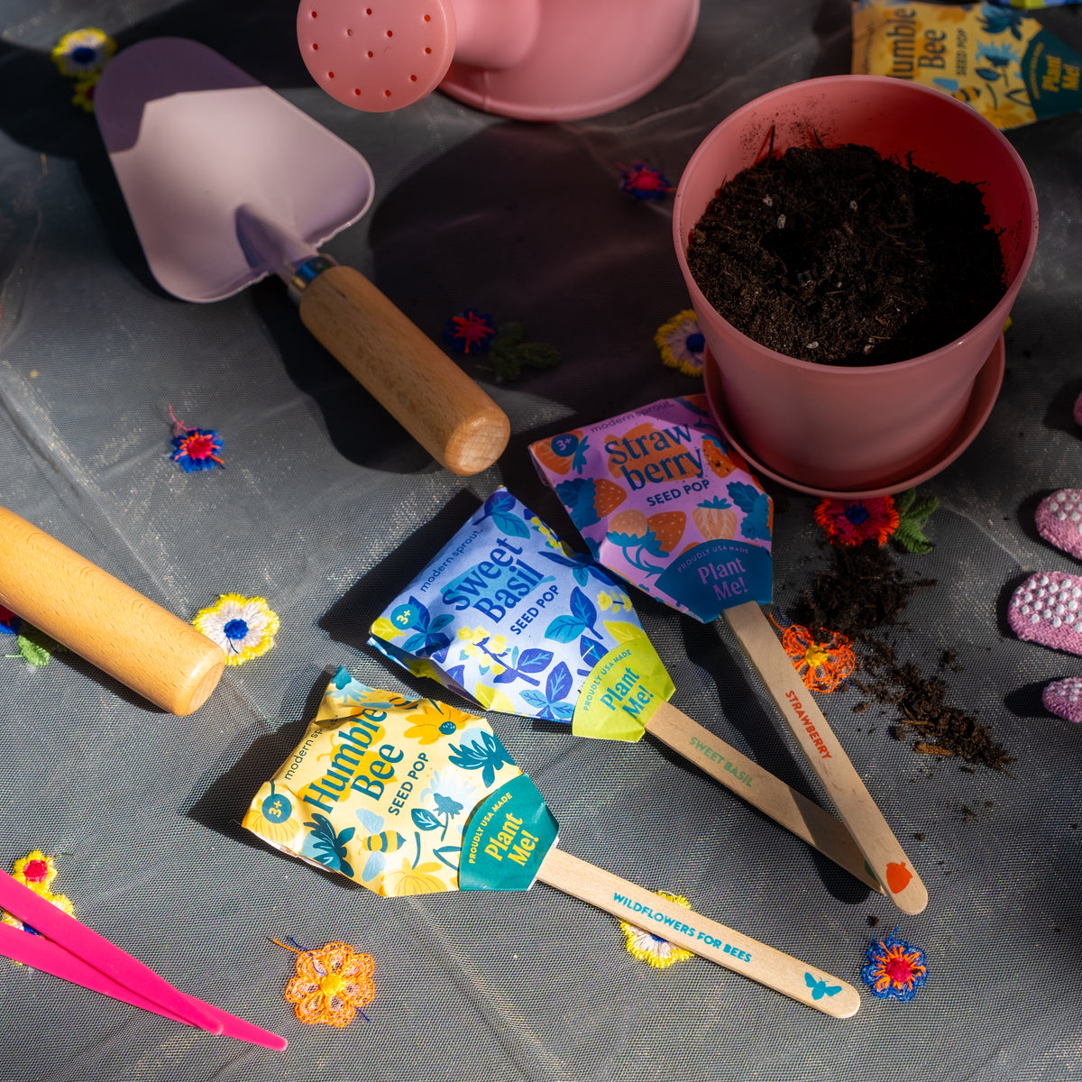 Seed Pops (Strawberry, Sweet Basil, Humble Bee), gardening tools, soil, a pink watering can, and plant markers sit on a floral cloth-covered table in bright sunlight.