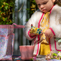 A young child in a yellow dress pours Seed Pops (Strawberry, Sweet Basil, Humble Bee) from a colorful packet into a pink flowerpot, with a floral bag and gardening kit parts scattered on the table nearby.