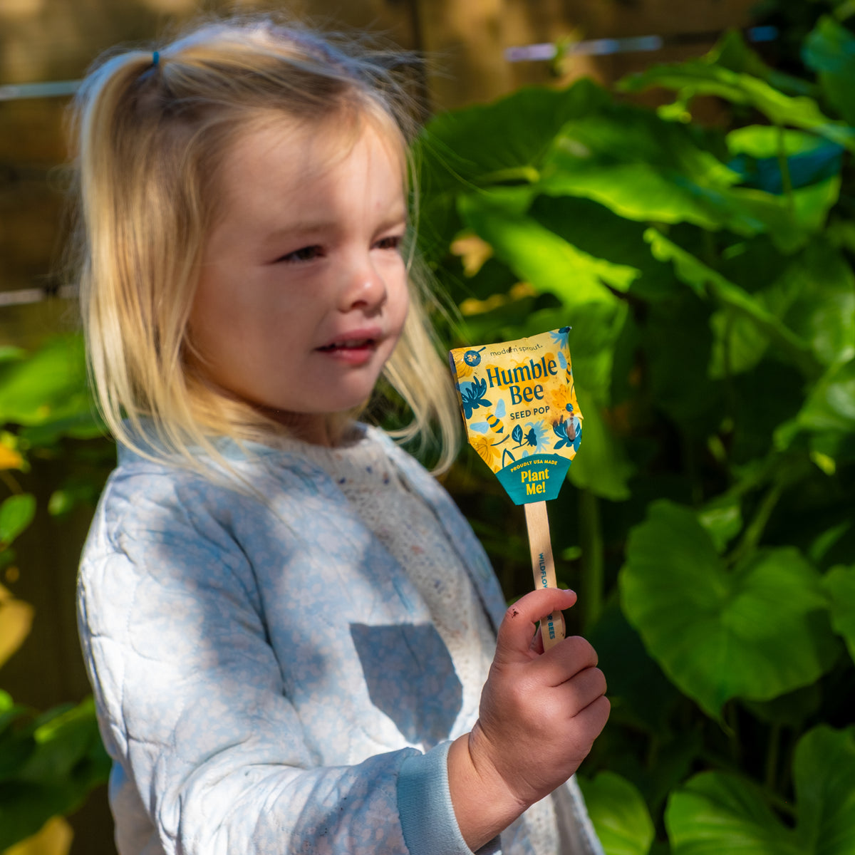 A young girl holds a Seed Pops (Strawberry, Sweet Basil, Humble Bee) gardening kit on a stick outdoors, sunlight shining and green plants behind her, ready to plant non-GMO seeds.