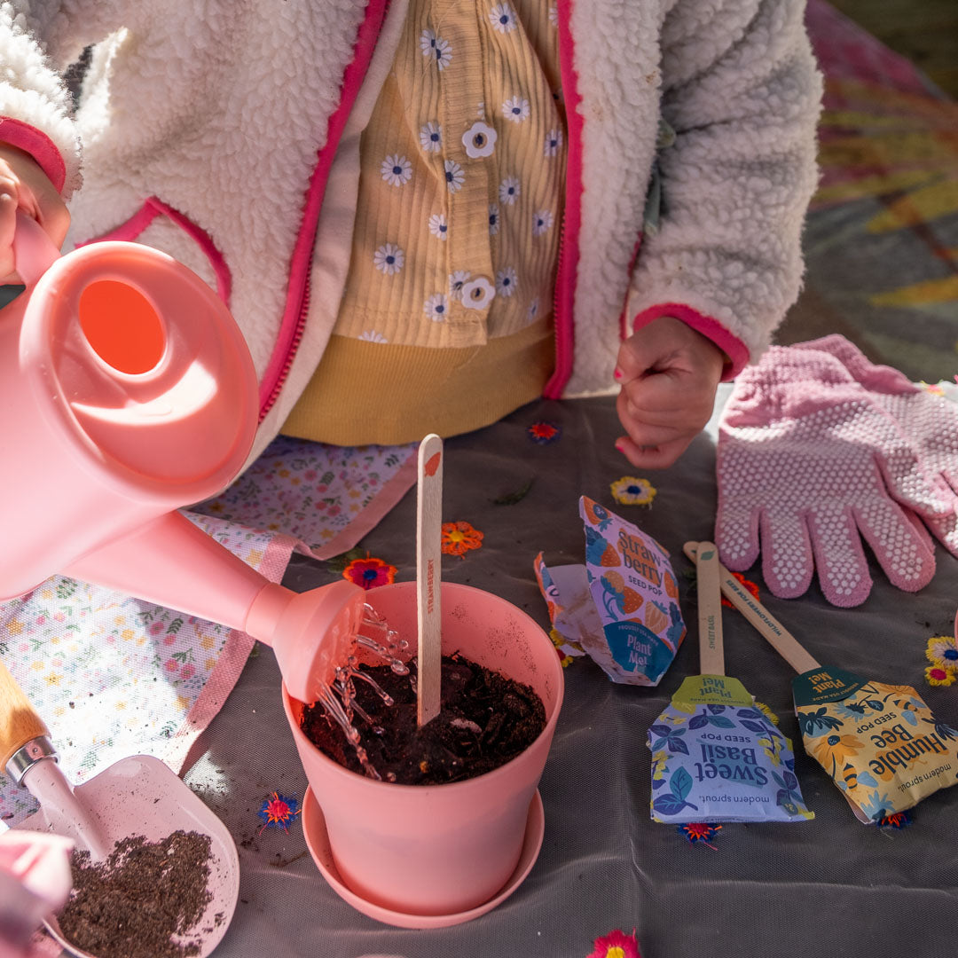 Children's gardening activity with a pink pot, watering can, and tools on a table.