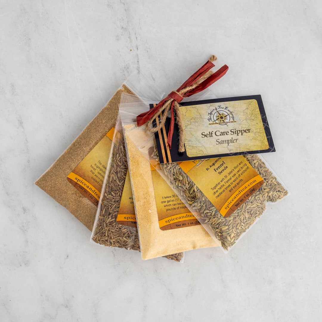 The Self Care Sipper Sampler features four clear pouches of coriander, cumin, fennel tea, and spices with yellow labels, tied with a red ribbon and tag—perfect for your daily ritual and elegantly arranged on marble.