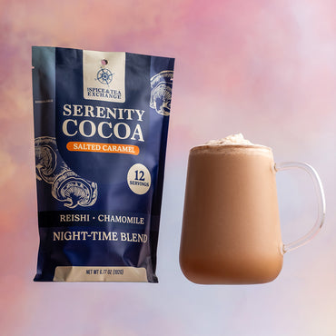 A bag of Serenity Cocoa Salted Caramel sits beside a glass mug of frothy cocoa, set against a soft pastel pink and purple background.