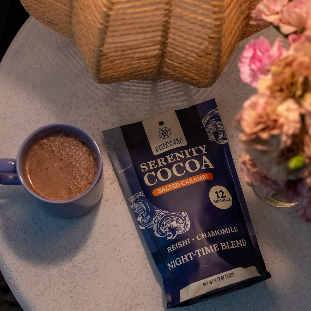 A packet of Serenity Cocoa Salted Caramel and a mug of evening cocoa rest on a round table by a flower vase and wicker lampshade, creating a cozy bedtime ritual atmosphere.