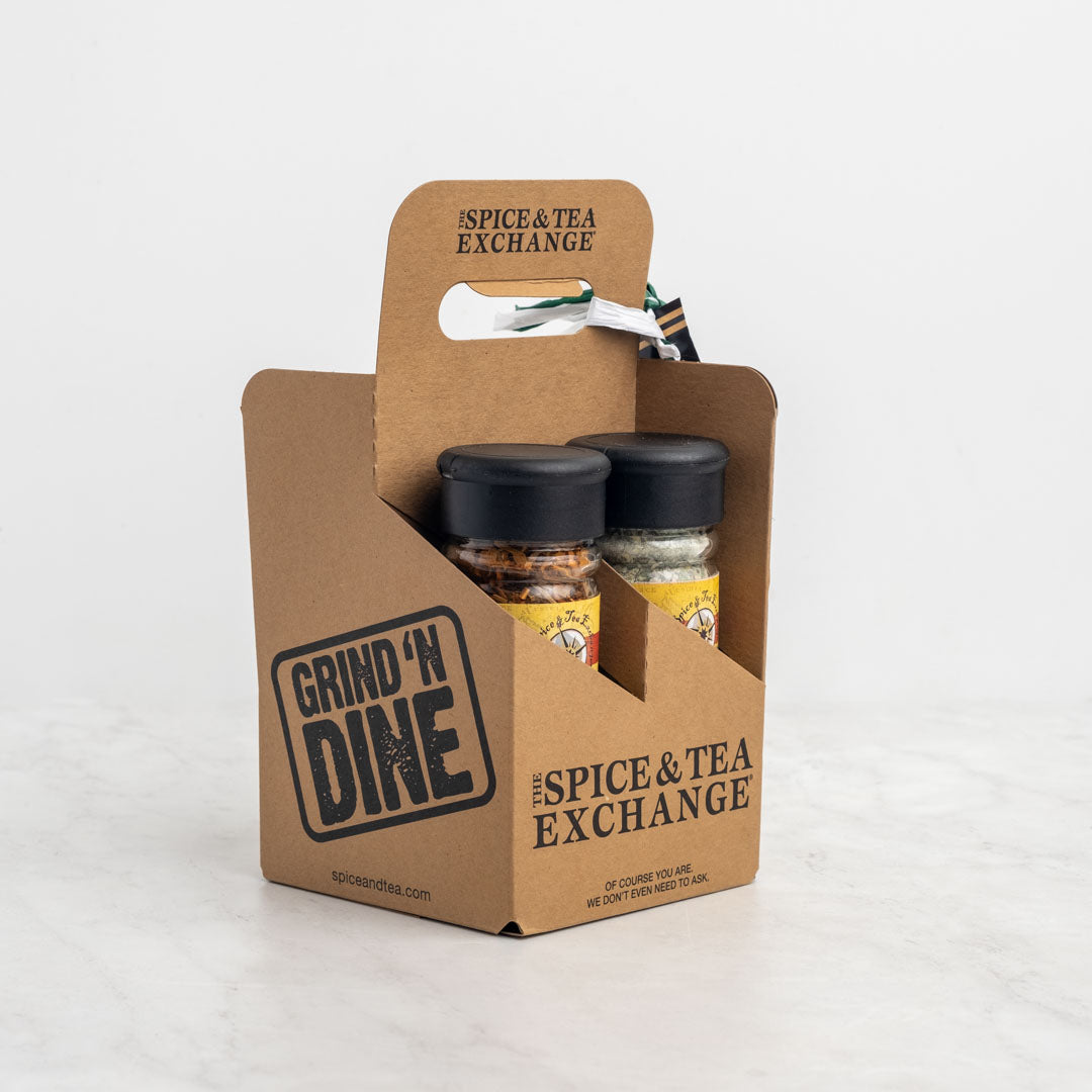 The Seriously Seafood 4-Pack Gift Set features four spice blends in a brown carrier labeled GRIND N DINE and The Spice & Tea Exchange, perfect for seafood dishes. The carrier has a handle, striped ribbon, and sits on a white surface.