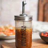 The Shake Strain & Pour Jar, featuring a metal pour spout and The Spice & Tea Exchange label, is filled with brown vinaigrette dressing and rests on a wooden surface with blurred dishes in the background.