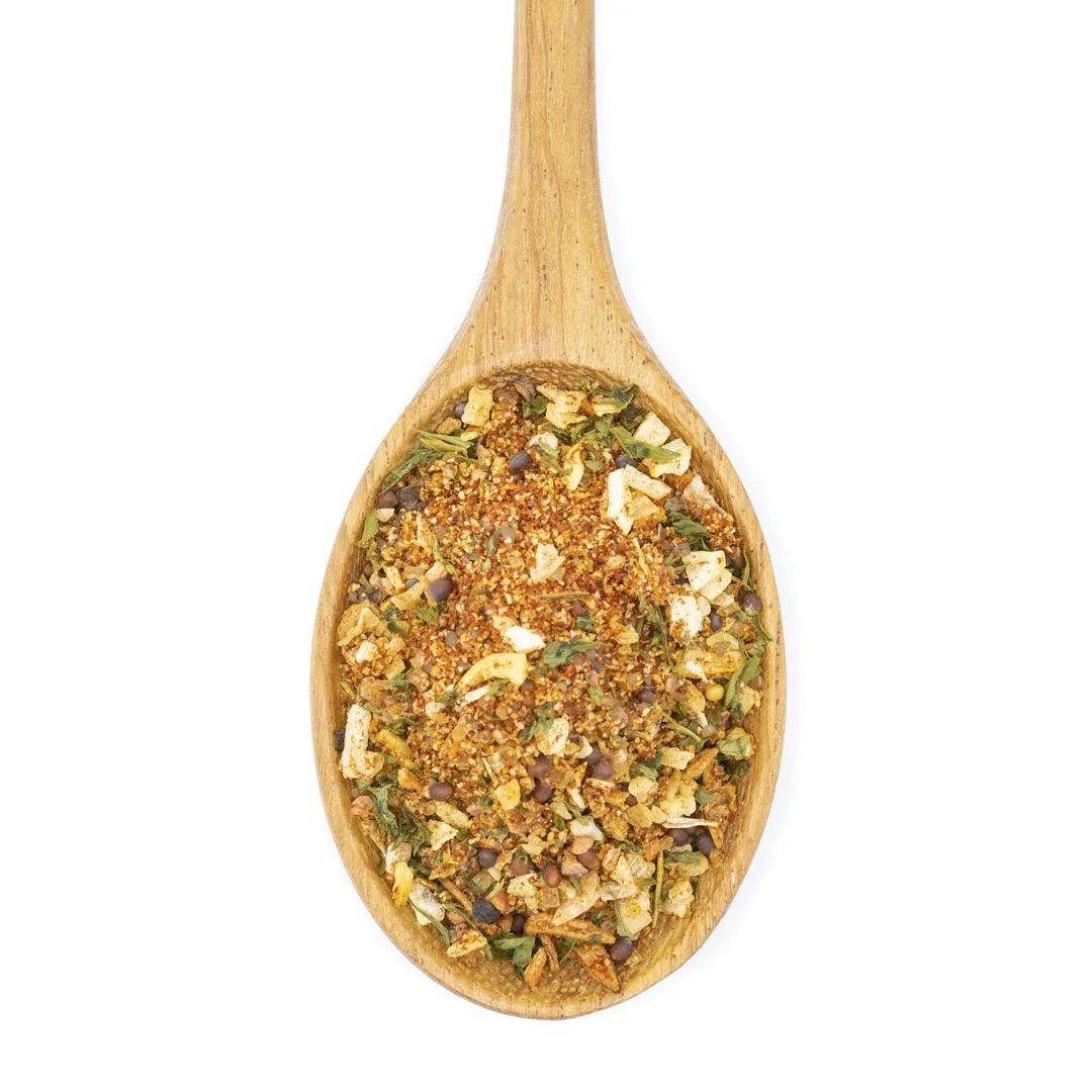 A wooden spoon filled with Smokehouse Rub Spice Blend—a mix of herbs, spices, chopped vegetables, and smoky sea salt—is displayed on a white background.