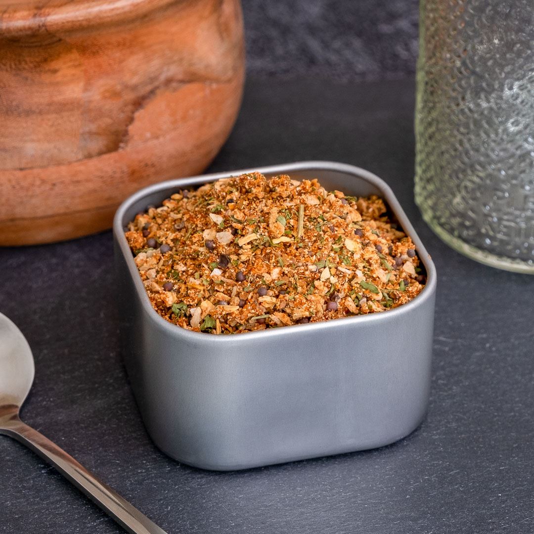 A square gray container of Smokehouse Rub Spice Blend sits on a dark surface beside a silver spoon, wooden bowl, and glass jar—ideal for bringing bold Midwest barbecue flavor to your next meal.