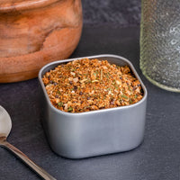 A square gray container of Smokehouse Rub Spice Blend sits on a dark surface beside a silver spoon, wooden bowl, and glass jar—ideal for bringing bold Midwest barbecue flavor to your next meal.