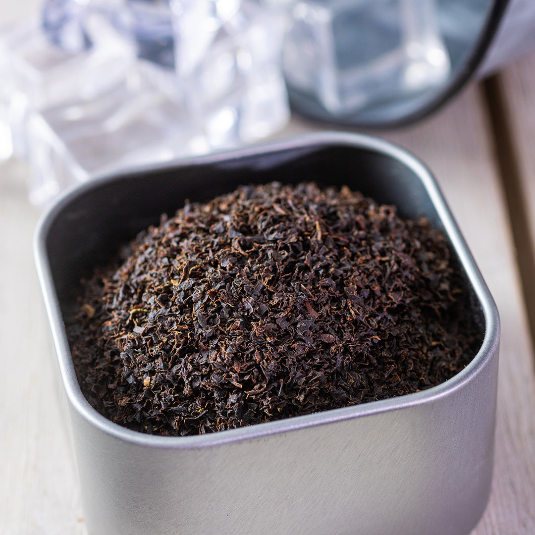 A square metal container of Southern Iced Tea loose black tea sits on a light wooden surface, with ice cubes and a glass in the background—ideal for brewing authentic southern iced tea.