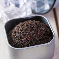 A square metal container of Southern Iced Tea loose black tea sits on a light wooden surface, with ice cubes and a glass in the background—ideal for brewing authentic southern iced tea.