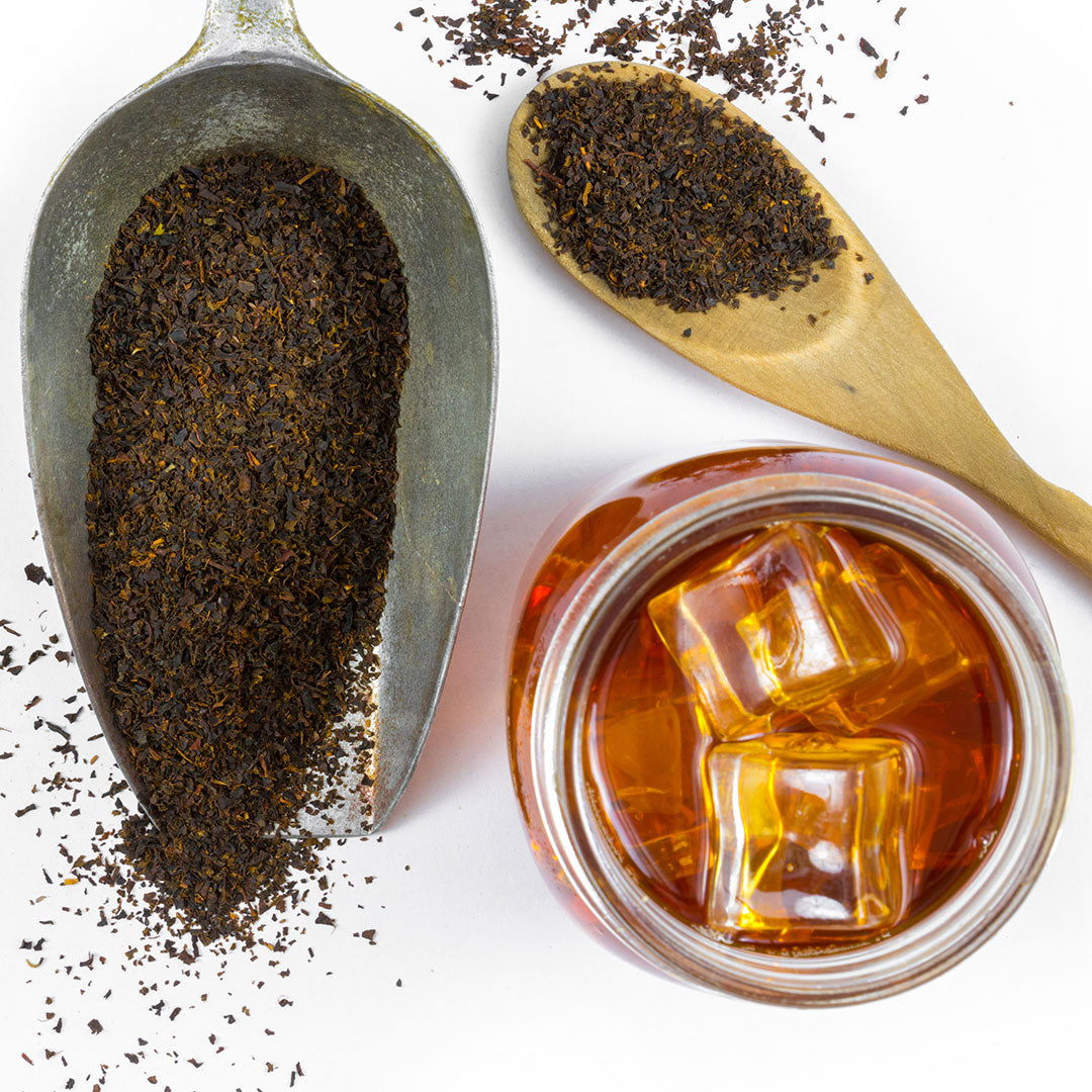 Southern Iced Tea is served in a glass with ice cubes, alongside loose black tea leaves displayed in a metal scoop and wooden spoon, all arranged on a white surface.