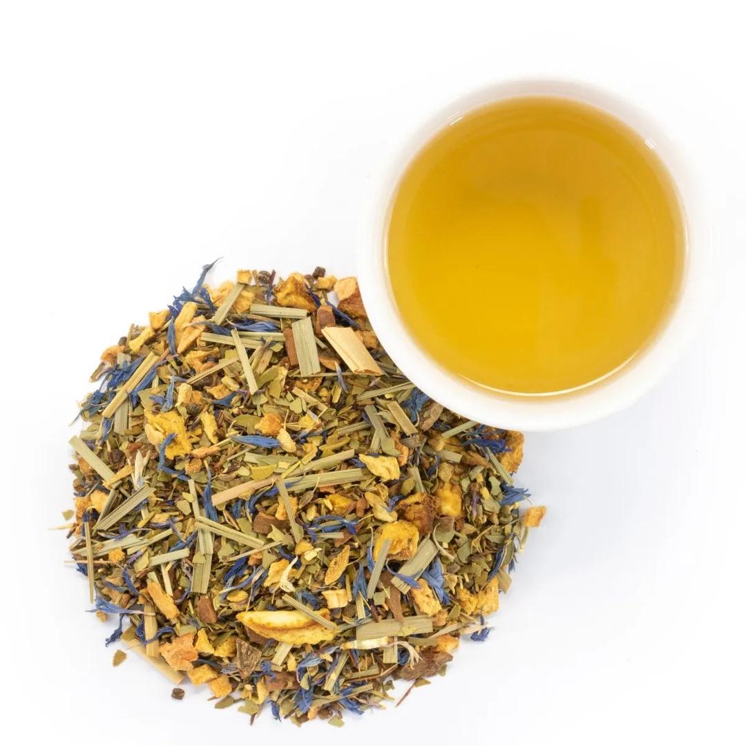 A white cup of Spiced Chai Mate Tea, light yellow in color, sits beside a pile of loose leaf blend with dried herbs, flowers, and fruit pieces on a white background.