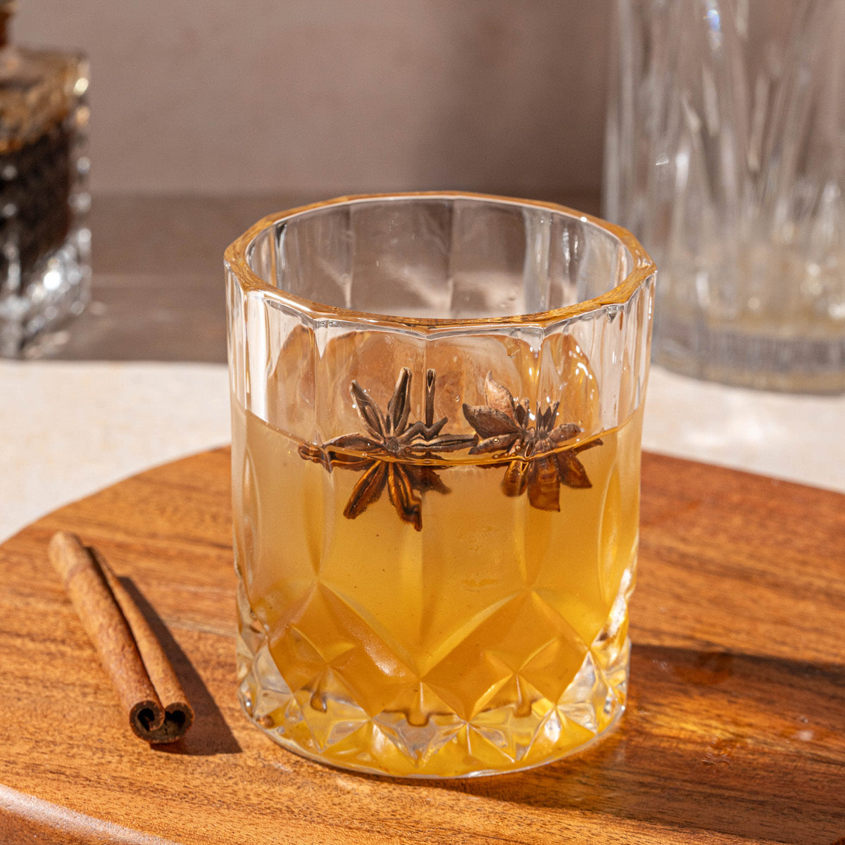 A glass of Spiced Chai Old Fashioned (Kit), garnished with star anise, sits on a wooden surface next to a cinnamon stick, with a blurred glass and seasoning kit in the background.
