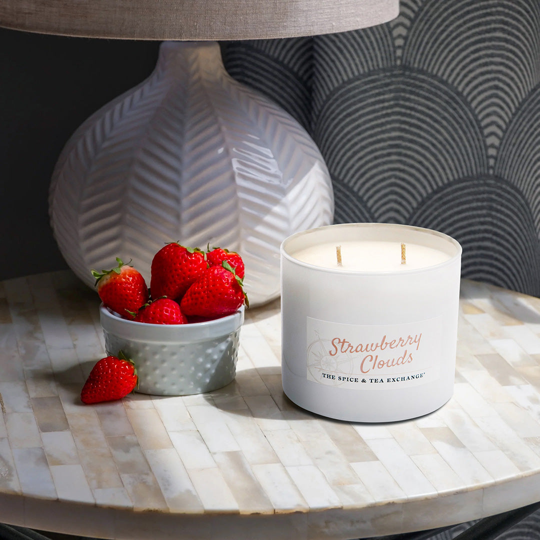 Candle labeled 'Strawberry Clouds' next to a bowl of strawberries on a table.