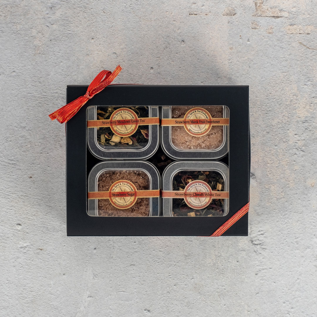 The Strawberry Fields 4-Tin Gift Box features four labeled clear tea tins, including strawberry tea, arranged in a black box with brown ribbons and topped with a red-orange ribbon, displayed on a light gray textured surface.