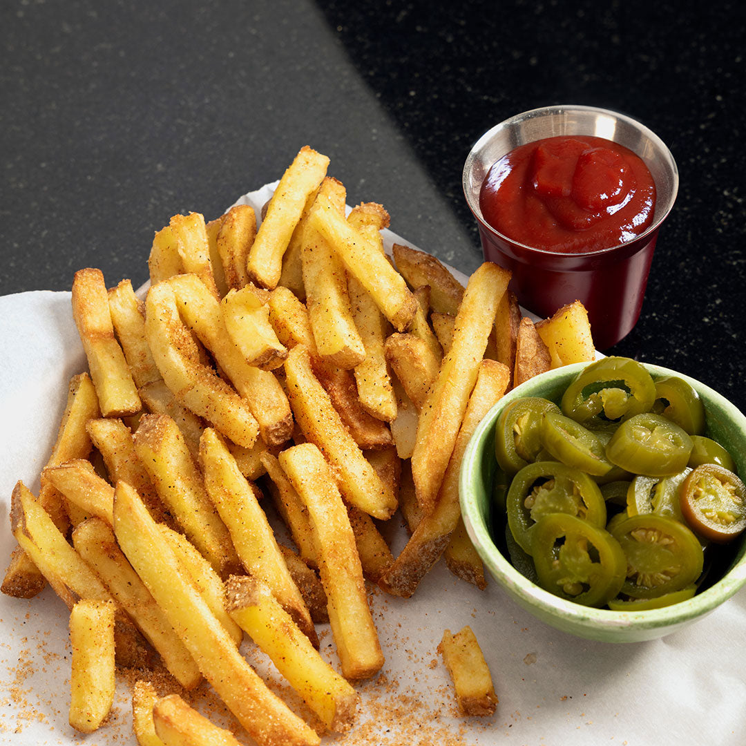 A pile of French fries tossed in Sweet 'n Smoky French Fry Seasoning is served on white paper, accompanied by a small metal cup of ketchup and a green bowl of sliced pickled jalapeños on a dark countertop.