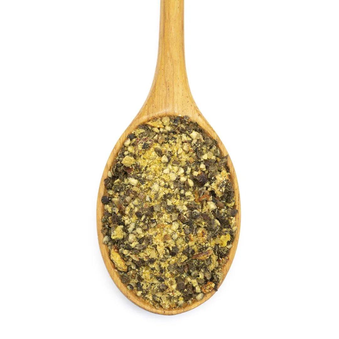 A wooden spoon holds Select Steak Spice Blend, a coarse yellow and green seasoning rich in flavor, set against a white background.