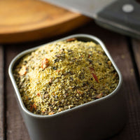 A square metal container of Select Steak Spice Blend, filled with a low-sodium mix of dried herbs and spices. A cutting board and knife are blurred in the background on wood, suggesting bold flavor.
