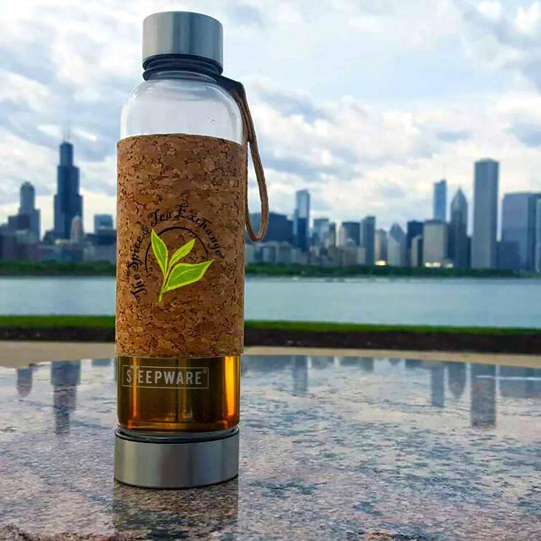 The Urban Tea Tumbler, featuring an eco-friendly design and a cork sleeve, sits on stone with the Chicago skyline and waterfront in the background beneath a cloudy sky, highlighting its modern style.