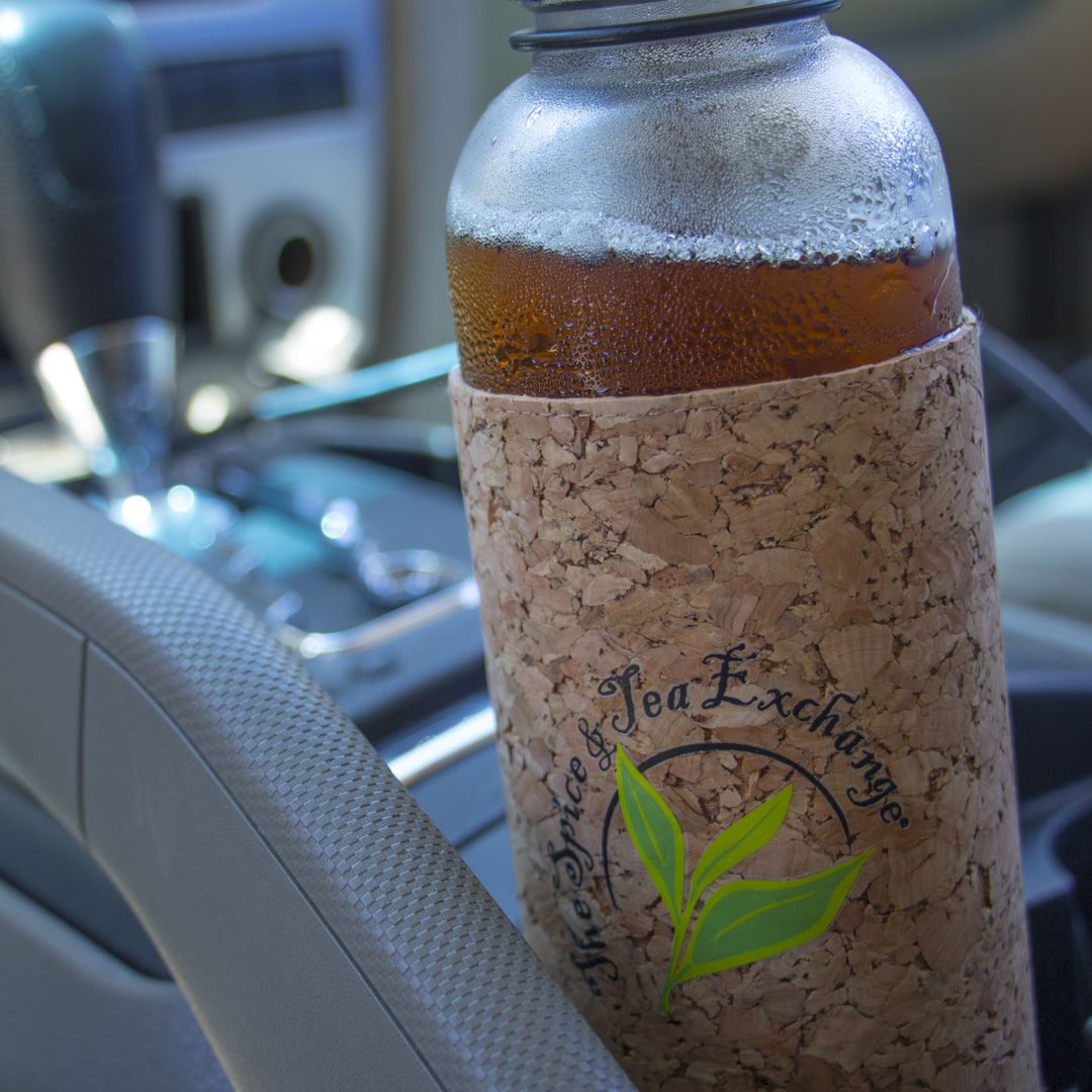 An Urban Tea Tumbler with a cork sleeve featuring The Spice & Tea Exchange logo and a green leaf design sits in a car cup holder. Condensation is visible on the eco-friendly glass bottle, with the car interior in the background.
