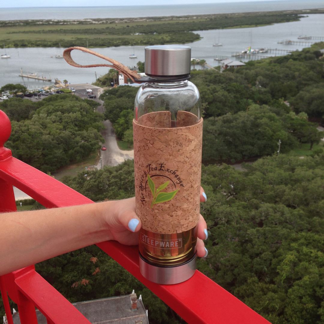 A hand holds the Urban Tea Tumbler with a cork sleeve and metal lids on a red railing, overlooking a river with trees and boats—a perfect eco-friendly companion for tea lovers.