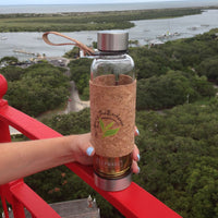 A hand holds the Urban Tea Tumbler with a cork sleeve and metal lids on a red railing, overlooking a river with trees and boats—a perfect eco-friendly companion for tea lovers.