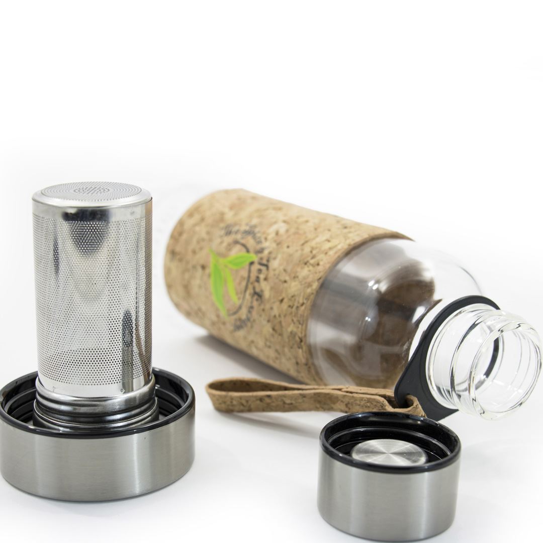 The Urban Tea Tumbler, featuring a cork sleeve and carrying strap, rests on its side. In front are its eco-friendly tea infuser and two stainless steel lids—one on the infuser, the other lying separately.