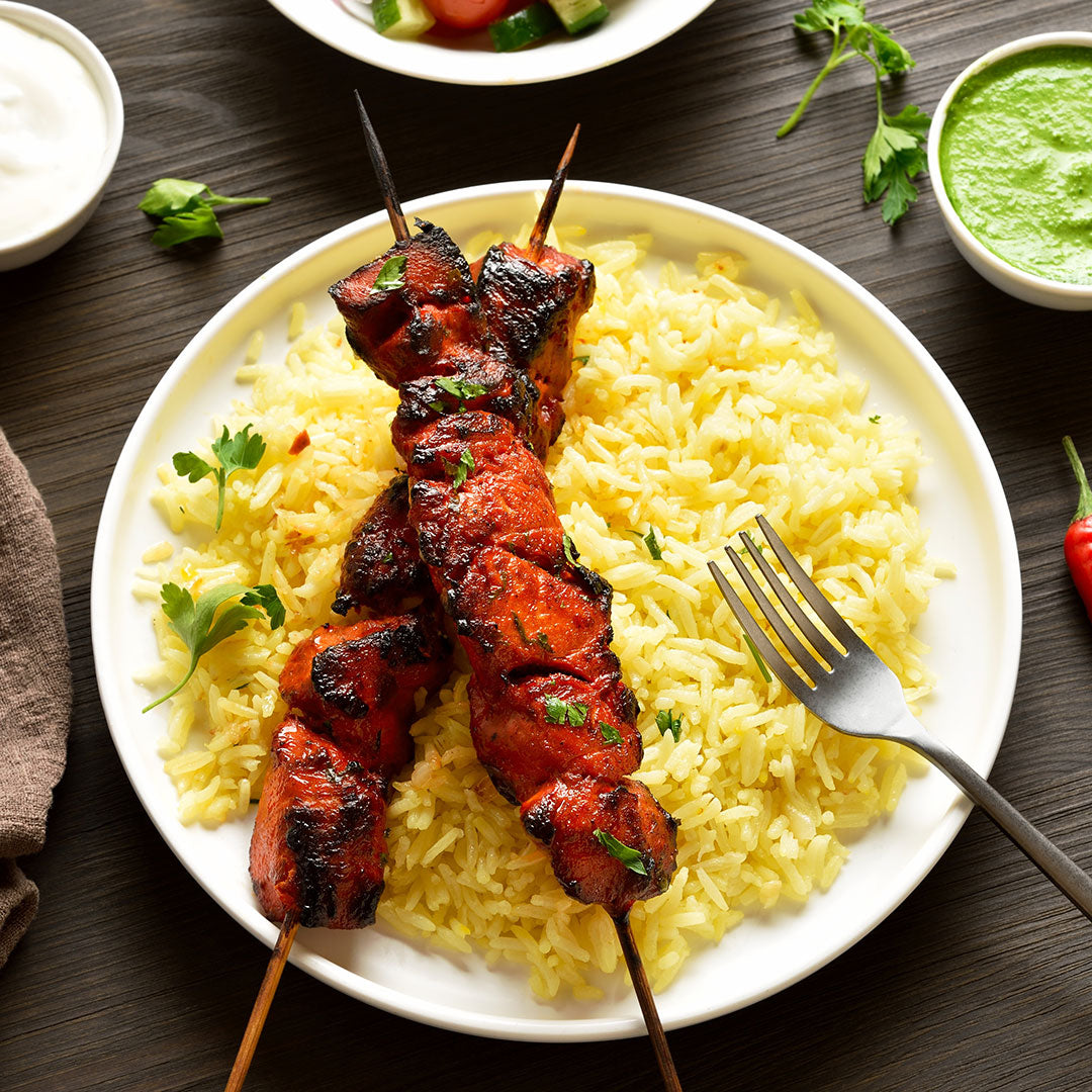 Skewers of grilled tandoori meat on a bed of yellow rice with a fork, on a wooden table.