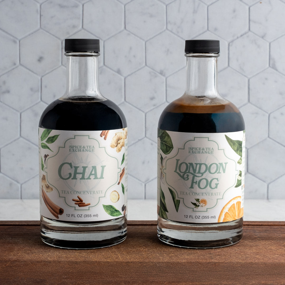 Two bottles of Spiced Tea Exchange Chai and London Fog on a wooden surface with a tiled wall background.
