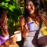 Two women enjoying tea in mugs outdoors with greenery in the background