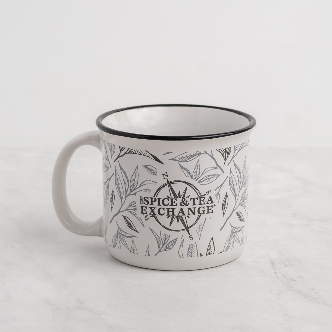 The Tea Leaf Ceramic Camp Mug is a 12-ounce white ceramic mug featuring a black tea leaf motif and "The Spice & Tea Exchange" printed in the center, shown on a light marble surface with a plain background.