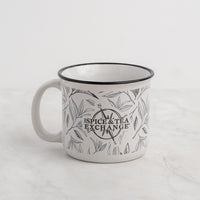 The Tea Leaf Ceramic Camp Mug is a 12-ounce white ceramic mug featuring a black tea leaf motif and "The Spice & Tea Exchange" printed in the center, shown on a light marble surface with a plain background.