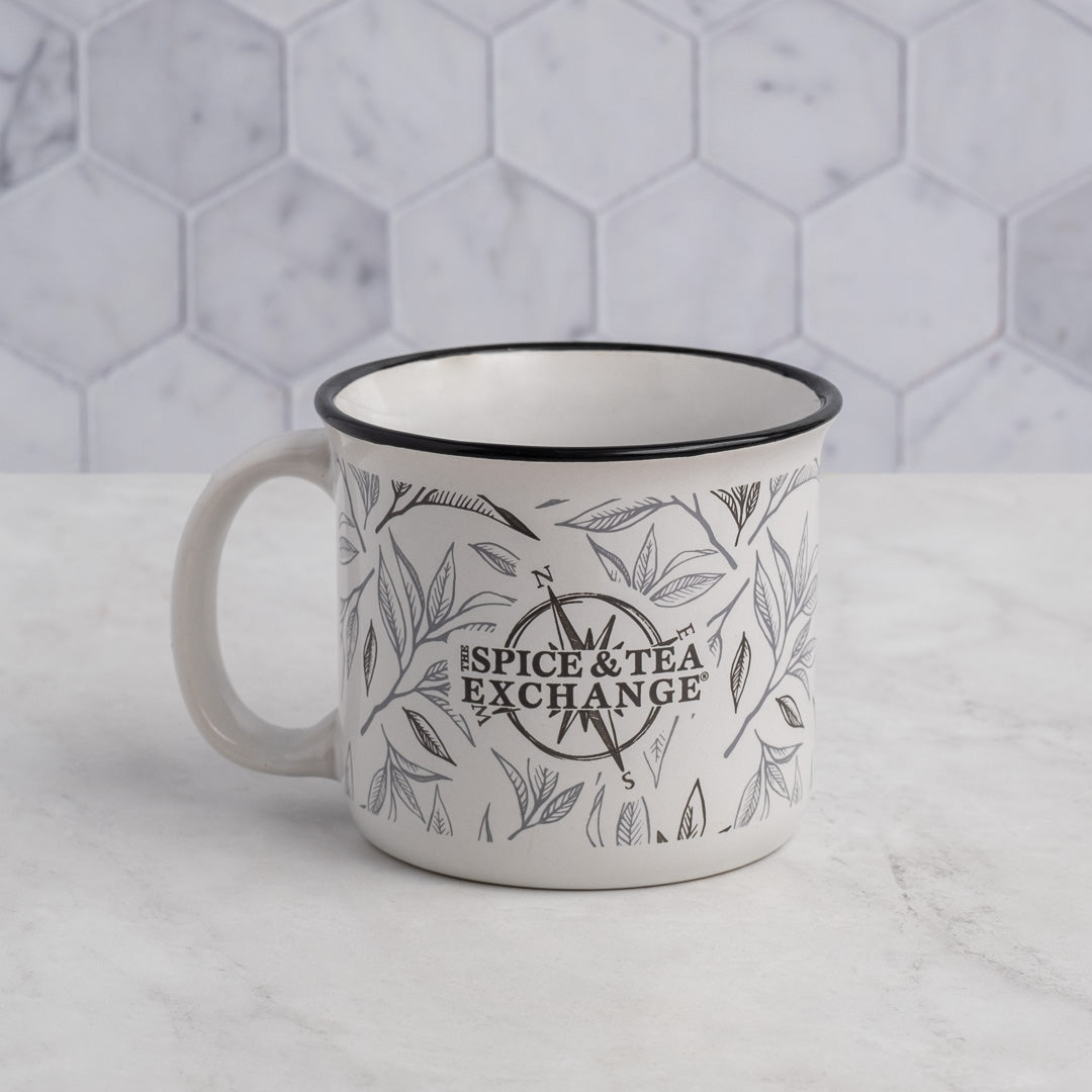 The Tea Leaf Ceramic Camp Mug, a 12-ounce mug featuring black tea leaf designs and The Spice & Tea Exchange text, rests on a light marble surface with a hexagonal white and gray tiled background.