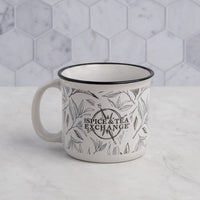 The Tea Leaf Ceramic Camp Mug, a 12-ounce mug featuring black tea leaf designs and The Spice & Tea Exchange text, rests on a light marble surface with a hexagonal white and gray tiled background.