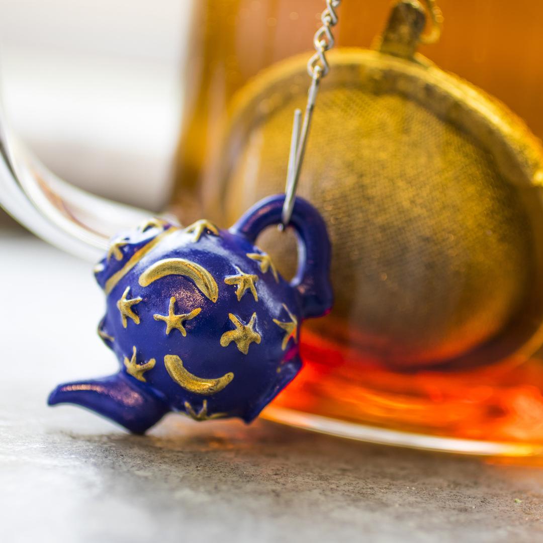 The Infuser with Teapot Charm is a blue, teapot-shaped loose leaf tea infuser adorned with gold stars and crescent moons, shown hanging in front of a cup of amber tea—a whimsical and charming travel accessory for tea lovers.