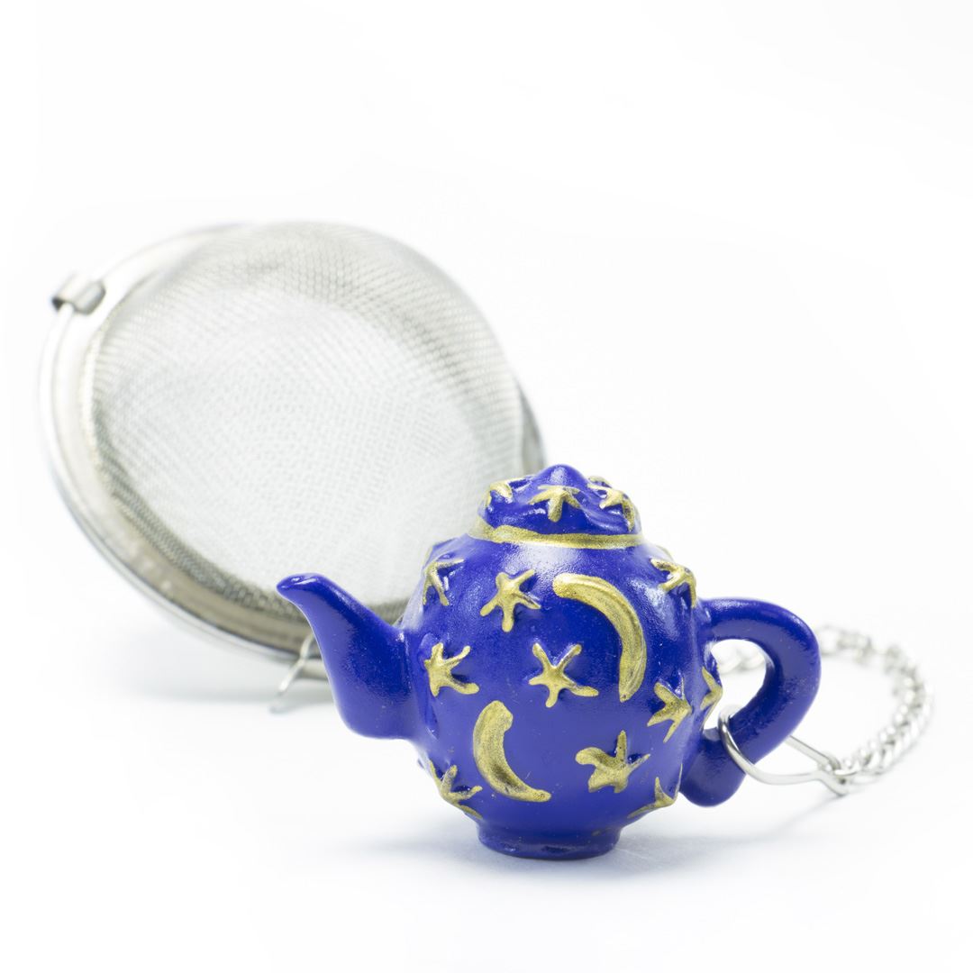 The Infuser with Teapot Charm features a silver tea infuser ball and a blue teapot charm adorned with gold stars and moons—a whimsical accessory ideal for brewing loose leaf tea and enhancing any tea lover's travel collection.