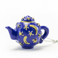 The Infuser with Teapot Charm is a small, dark blue teapot keychain adorned with gold crescent moons and stars, making an enchanting travel accessory for tea lovers. Displayed against a plain white background.