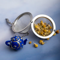A charming blue teapot adorned with gold stars and moons sits beside the Infuser with Teapot Charm, filled with dried chamomile flowers, some scattered across a blue and white surface.